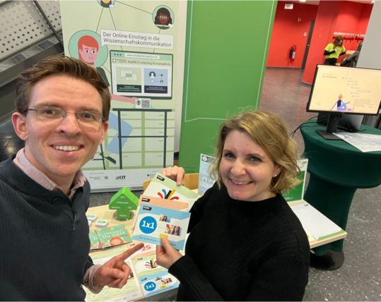 Der NaWik-Messestand auf der DPG-Frühjahrstagung am KIT in Karlsruhe. Zwei NaWik-Mitarbeitende stehen vor dem Messestand und verweisen auf die Angebote.