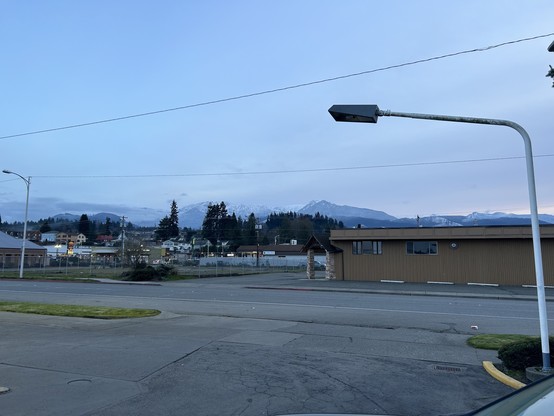 Snowy mountains in the distance under a mostly clear sky, with a street in the foreground, a single-story building to the right, power lines and a streetlight above. 
