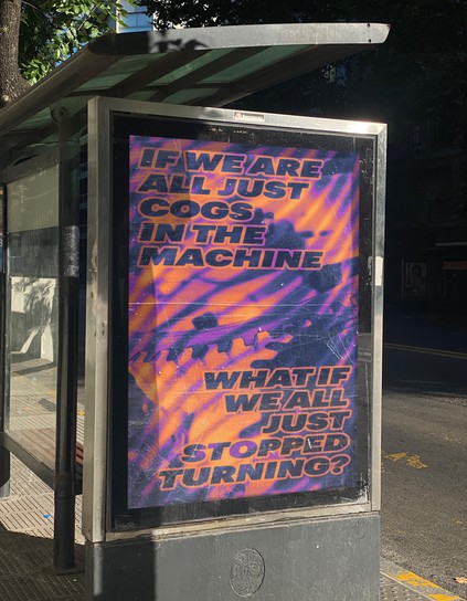 image of poster in bus shelter advertisement frame with scuffed and worn plexiglass on a sunny day. the road runs diagonally through the image so the poster is seen at a slight angle and partially shaded. the poster shows heat-map like imagery (purples, magentas, oranges blended in gradients) of closeups of cogs. in an oblique, wide, sans serif font in all caps and dark purple with an orange stroke, the text reads “IF WE ARE ALL JUST COGS IN THE MACHINE WHAT IF WE ALL JUST STOPPED TURNING?”