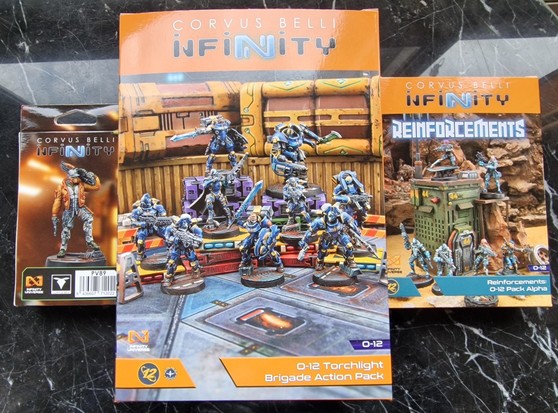 La imagen muestra tres cajas de Infinity por el lado de las miniaturas, de izquierda a derecha: nuevo cazarrecompensas de la Adepticon 2024, Action pack de la brigada Torchlight y pack alfa de refuerzos de O-12