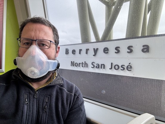 A man sitting on a BART light rail train at the Berryessa station in San Jose, California. The man is wearing glasses and a personal air purifying respirator (PAPR), for pandemic safety reasons.