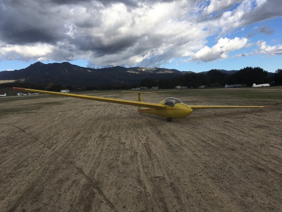 A yellow glider sits on a dirt runway. In the background, mountains rise below dramatic clouds