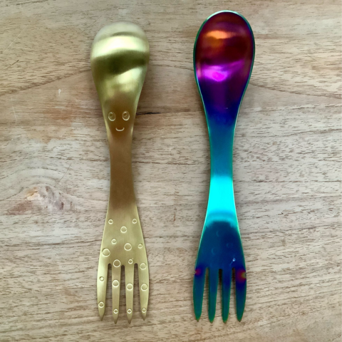A golden spork and a rainbow spork shown on a wooden background.