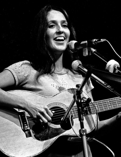 Joan Báez, pelo largo oscuro y guitarra al frente, sonríe mientras canta sobre el escenario, en una fotografía en blanco y negro de 1973. Heinrich Klaffs • CC BY-SA 2.0
