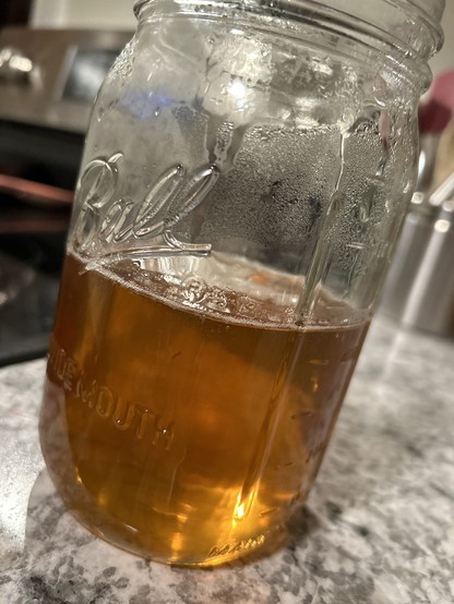 A large ball jar, a mason jar, is sitting on fake granite countertop. It’s filled 1/3 of the way with a golden brownish liquid, it’s maple syrup!