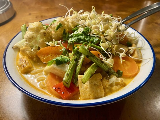Large blue rimmed white bowl of a creamy red soup feat submerged noodles, tofu and carrots, piled high with slightly blackened pan fried veggies (baby corn, green beans, cauliflower, peppers, carrot) and topped with lime juice, lentil sprouts and coriander