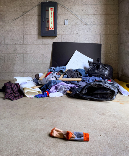 Color photo of a pile of unfolded clothing on a concrete floor in front of a cinder block wall. Attached to the wall is a fire extinguisher enclosure. Leaning against the wall is a black piece of poster board with a smaller, white poster board in front. In the foreground is a solitary multi-hued sock.