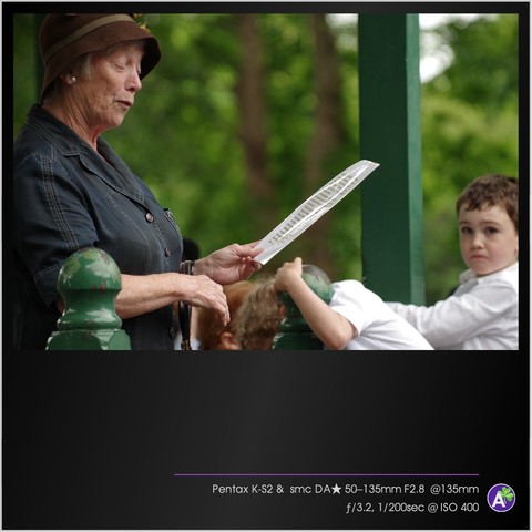 a color outdoor photograph. an older woman stands on a porch or in a gazebo. she is reading from a laminated page. her face appears animated and energetic. several people listen in the background. two young children, a boy and a girl, look very bored. the boy climbs a pole while the girl hides her head in her arms. there are no animals or vehicles in this picture.