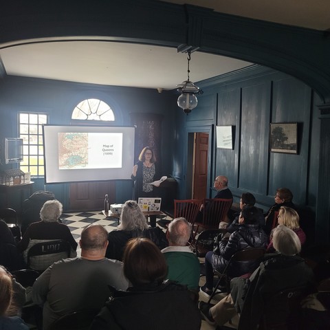 dimly lit photo of audience listening to me talk about Naming Gotham. I am showing them an 1899 map of Queens.  sigh.  Beautiful 17th century hall with paneling and half-windows barely visible. 