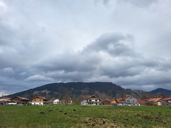 This image captures a serene, picturesque landscape embodying the essence of rural tranquility. A cluster of houses, modest in structure and possibly featuring red roofs, nestles within a vast field that stretches towards the horizon. The field, rich with lush green grass, hints at the fertility of the land and the harmony of living close to nature. In the background, a hill rises gently, its presence adding a sense of majestic quietude to the scene. Above, the sky is a canvas of drama and calm…