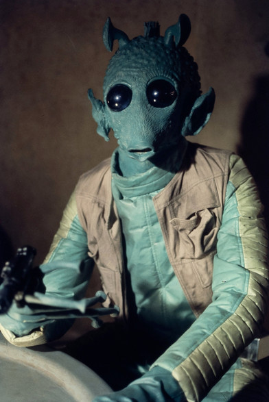 Greedo from Star Wars sitting in the Mos Eisley cantina holding his blaster.