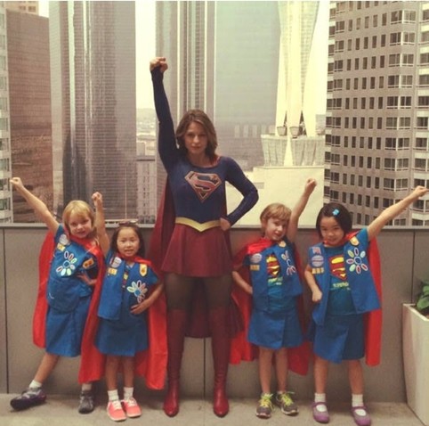 Photo of Melissa Benoist as Supergirl, with one hand on her hip and the other fist held high, as if she is preparing to fly. She is surrounded by four Daisy Girl Scouts, all wearing matching blue skirts and badge-laden Daisy vests and red capes, and all of whom are affecting the same pose. At least two of the girls have Supergirl's "S-shield" on their own chests.