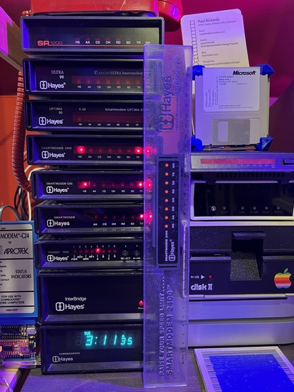 A stack of Hayes modems of various speeds and other peripherals. A Hayes tiller stands next to it, along side an Applesauce, a Disk II floppy drive, and a lone USR Courier modem. 