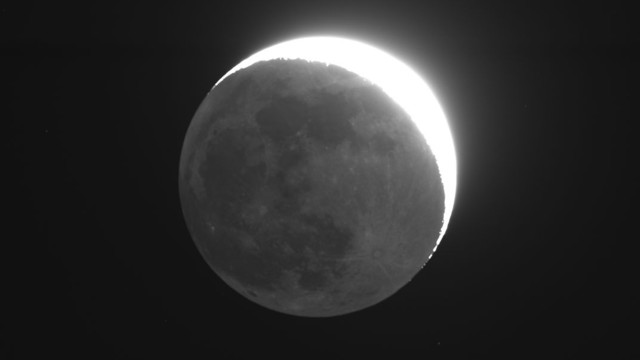 Black background. The moon fills most of the image. A thin crescent part of the moon (upper right) is very bright white and over-exposed. The rest of the moon is dim but surface details discernable (darker "Mares" and brighter craters).