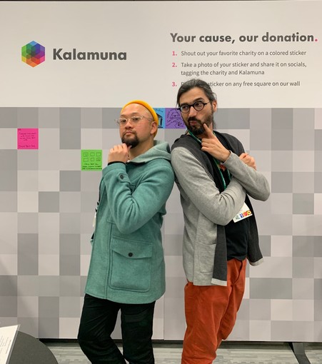 Ken and Andrew stand in front of the booth wall with hands on their chins and look as if they're pondering something. The wall has different shades of grey squares, with several stickers already placed.