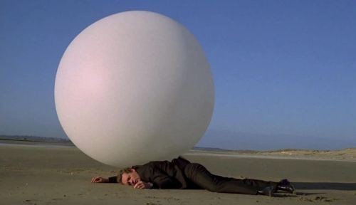 Rover - a giant white ball - sits atop a prone Patrick McGoohan (Number 6) on the beach in the show The Prisoner.