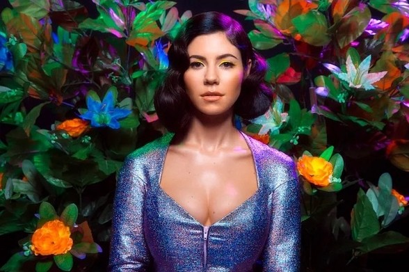 Marina Diamandis con un vestido de latex con textura glitter, sobre un muro de flores con colores neon brillantes.