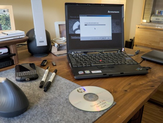 Photo showing my Thinkpad X61 with its factory restore recovery progress on-screen. In front of the Thinkpad is one of the burned discs used to start this restore process. To the left of the laptop are two felt markers and some USB sticks, along with my Unihertz Titan Pocket phone that looks like a Blackberry with a hardware keyboard. On the right in the background you can spot part of a European Super Nintendo standing in a cabinet. All this put together kind of give this photo a strong retro vibe, as if it could've been taken sometime in the mid-2000s.