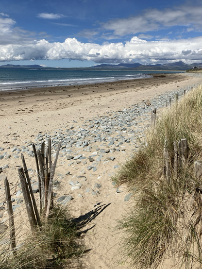 The end of a path through the dunes, descending onto an empty beach. It’s stony nearer the dunes, then sandy (the sand is darker and damp near the sea; the tide is clearly going out). The sea is a deep jade / peacock colour; the sky blue but with clouds across it.