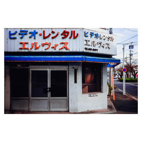 The photo depicts an old video rental shop with faded signage that reads "Elvis" in bright colors above the entrance. The shop's exterior is simple and a bit worn, hinting at the countless stories it has seen over the years. The street is quiet, and the shop seems to be waiting for visitors to explore the cinematic treasures that might lie within its walls. The scene is a nostalgic nod to the era of video rentals, a once-vibrant industry now quieted by the passage of time.