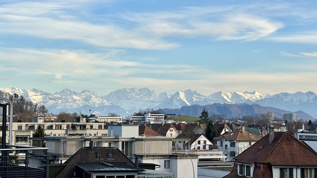 Die schneebedeckten Berner Alpen unter einem, mit ein paar Wolken verzierten, blauen Himmel. Im Vordergrund die Dächer Ostermundigens.