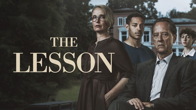 Julie Delpy, Richard E. Grant, Daryl McCormack und Stephen McMillan  stehen alle mit ernsten Mienen vor einem allen Herrenhaus im Garten, im Hintergrund ist ein in den Teich ragender Steg zu erkennen. Im Vordergrund der Schritzug in großen gelb-weißlichen Lettern "The Lessen"