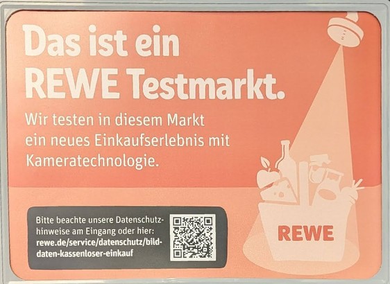 Foto eines roten Hinweisschildes im gerade eröffneten Rewe Hamburg, Hoheluftchaussee:  Rechts oben ist eine Art Duschtasse zu sehen, die aber eine Kamera sein soll und wie das Licht aus einem Ufo von oben einen gefüllten Einkaufskorb beleuchtet, der u.a. einen losen Apfel, eine Milchkanne, losen Käse zeigt, also einen klassischen Wochenmarkteinkauf, da diese unverpackten Waren in dem neuen REWE gar nicht erhältlich sind.
"Das ist ein REWE Testmarkt. Wir testen in diesem Markt ein neues Einkaufserlebnis mit Kameratechnologie." weiter unten in kleinerer Schrift: "Bitte beachte unsere Datenschutzhinweise am Eingang oder hier: https://www.rewe.de/service/datenschutz/bilddaten-kassenloser-einkauf dazu der passende QR-code. 
