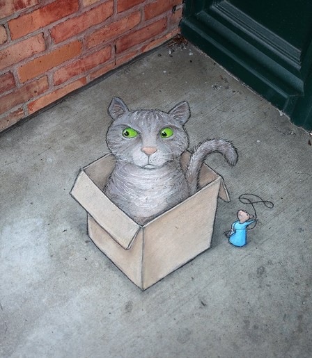 Street Art. Zeichnung auf dem Boden.
Eine graue Katze mit hellgrünen Augen sitzt in einem Karton. Daneben steht eine Maus, gekleidet in einem hellblauen Kleid, und schwingt ein Lasso gegen die Katze.
