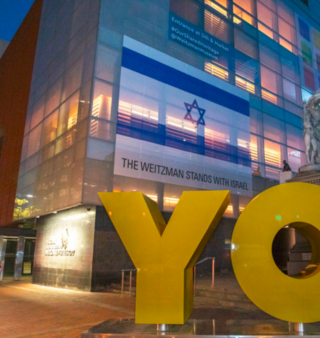 Photo: The corner of the Weitzman National Museum of American Jewish History in Philadelphia. There is a statue out front, spelling the word, "YO." There is an Israeli flag covering some of the illuminated windows of the museum. At the bottom it says, "The Weitzman stands with Israel."