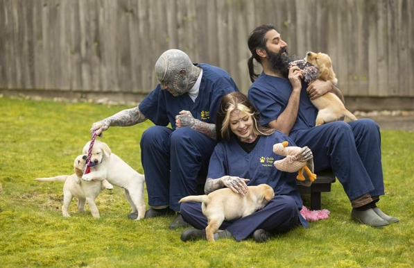 Someone with facial tattoos, someone with long hair and a beard, and someone with dyed hair and sleeve tattoos sitting down on grass, wearing blue scrubs and playing with cute Guide Dog puppies