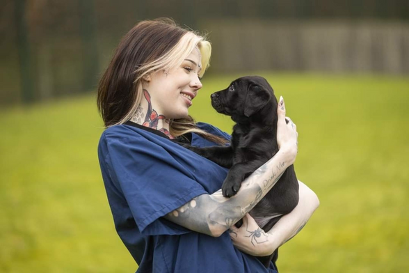 Someone with neck and arm tattoos and long brown hair with a blonde streak, smiling, holding a black lab puppy and looking at it