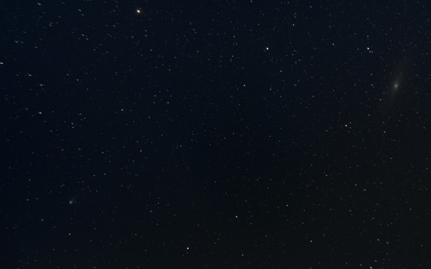 A colorful start field. At the upper right, the Andromeda Galaxy M31 is visible as an oblong reddish smudge, and at the lower left, a small bright green blob with a faint tail pointing toward the upper right is the comet