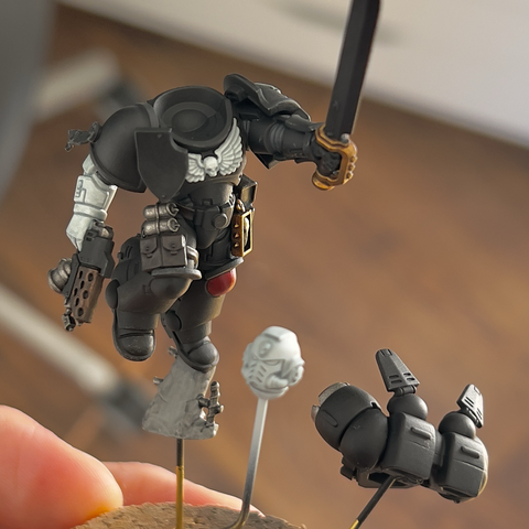 A Warhammer 40,000 miniture being painted. Various sub-assemblies are pictured on wires stuck into a cork base. 