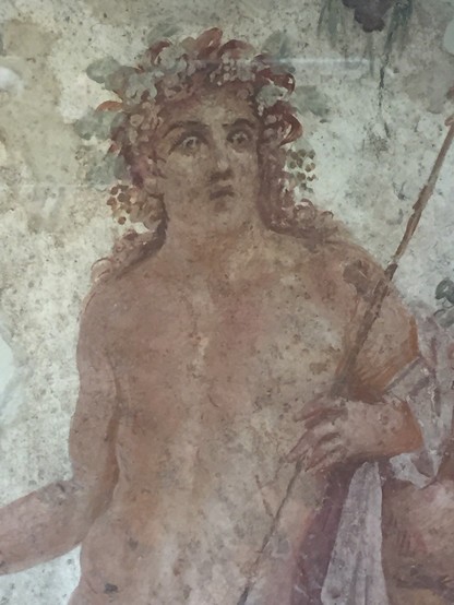 Fresco of Dionysos looking terrified. He has long brown hair with a grapevine crown and grapeclusters hanging down at his temples. He holds a thyrsos staff in his left and a piece of cloth is draped over the left arm of the otherwise nude god.