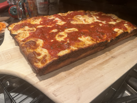 A side view of a Detroit pizza right out of the oven and pulled out of its pan ready for cutting. It is rectangular with visible cheese and rivers of tomato sauce cutting through the cheese, it’s on a wooden pizza paddle.