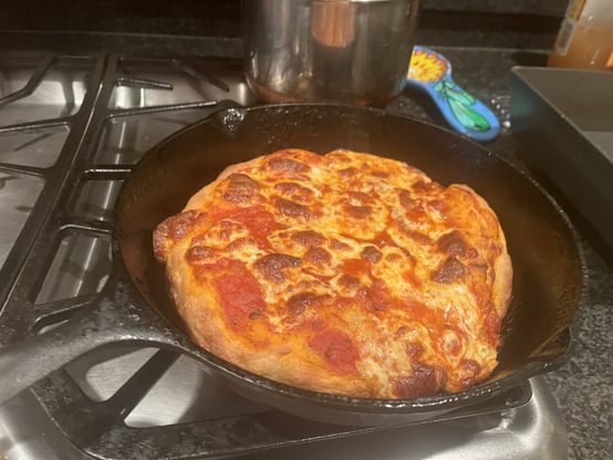 A cast-iron pan with a deep dish pizza in it, the pan is sitting on top of a stove with the pizza in it, it’s a gas stove with various other pans, and pots visible just toward the edge of this image