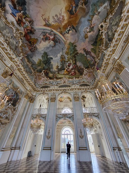 The image captures a serene and artistic atmosphere within an indoor setting, showcasing a man gracefully positioned amidst a room adorned with meticulously painted ceilings. The artistry above exudes an aura of historical elegance, possibly reflecting intricate patterns or storytelling murals that draw the viewer's gaze upward. Dominating the scene are shades of grey, both in the foreground and background, suggesting a harmonious blend of colors that enhances the aesthetic appeal without overp…