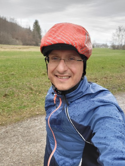 In this vividly captured frame, a man exudes joy as he beams with a genuine smile, donned in a protective helmet that hints at an adventurous spirit. The backdrop is a serene outdoor setting, where the lush green grass softly contrasts with the expansive sky above, suggesting a day perfect for hiking or perhaps a leisurely stroll in the park. His attire, a blue jacket, not only adds a pop of color to the scene but also suggests a readiness for varying weather conditions. Surrounding him, the na…