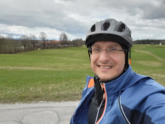 In this vibrant outdoor scene, we observe a man radiating joy with a broad smile on his face. He is adorned with a bicycle helmet, indicating a possible readiness for a cycling adventure. The backdrop is a picturesque setting that includes lush green grass that stretches across the landscape, hinting at a serene and open outdoor environment. Above, the sky is adorned with wispy clouds, suggesting a pleasant, clear day. The man's attire suggests he is prepared for physical activity, possibly cyc…