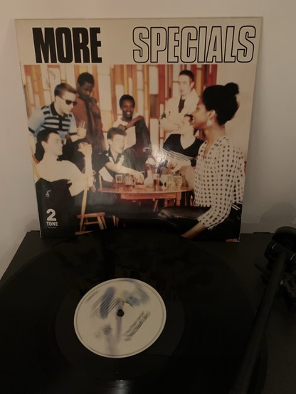 Foto einer Vinyl-Schallplatte auf einem Plattenspieler mit dem Albumcover "More Specials" von der Band The Specials im Hintergrund. Das Cover zeigt eine Gruppe von Menschen, die sich in einer Bar treffen.