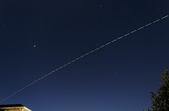 A composite image showing a late twilight sky, blue near the horizon fading to black at the top, the roof of a building the lower left and a tree in the lower right. Many stars are visible, perhaps as many as you'd see in a very dark location. ISS is visible as a dashed line, starting with short red dashes at the horizon on the lower left, increasing in length and fading to bright yellow-white as it passes over head. The brightest "star" in the scene, above the path of ISS is Jupiter. A slightly reddish smudge below and to the left is the Andromeda galaxy (M31) and a smaller green smudge below and to the left, only discernible if you enlarge the image is comet 12P Pons-Brooks
