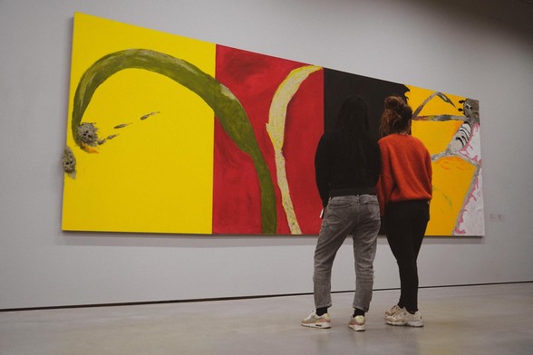 Two women stand before a large painting by Italian artist Mimmo Paladino. The painting consists of four large colour blocks next to each other: yellow, red, black and orange. On the yellow block, there are two protruding elements, which resemble faces. The red block features two organic shapes, one of which also overlaps the yellow block. The orange block contains a human-like figure.