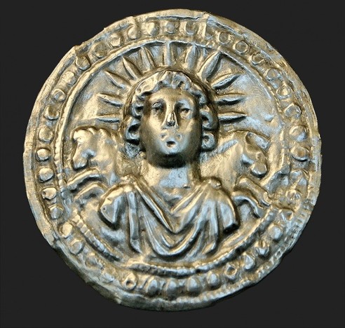 Silver disc with a relief of Sol Invictus. The god wears a sunray crown and a chiton or tunic pinned on his shoulders. Behind him, we see two of his prancing steeds, one to each side.