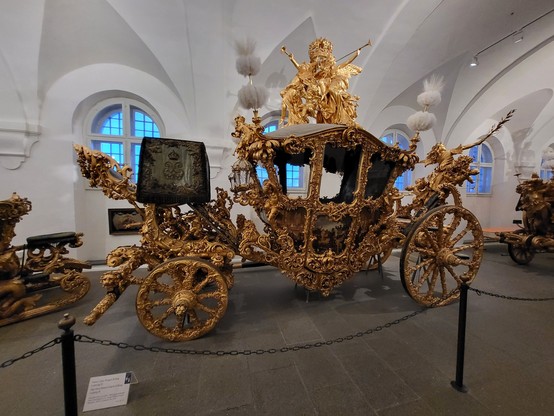 This image captures a regal and opulent scene, featuring a gold carriage that commands immediate attention. Atop the carriage, a statue adds a touch of classical elegance, suggesting a possible historical or commemorative significance. The carriage itself is intricately designed, with details that gleam in gold, indicating a level of craftsmanship that speaks of luxury and grandeur. Surrounding the carriage, the environment appears to be indoors, possibly within a museum setting, given the cont…