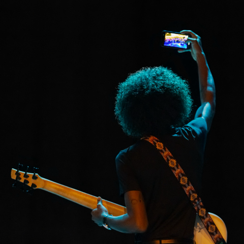 Chitarrista di colore con pettinatura afro fotografato di spalle mentre si fa un selfie con il cellulare.