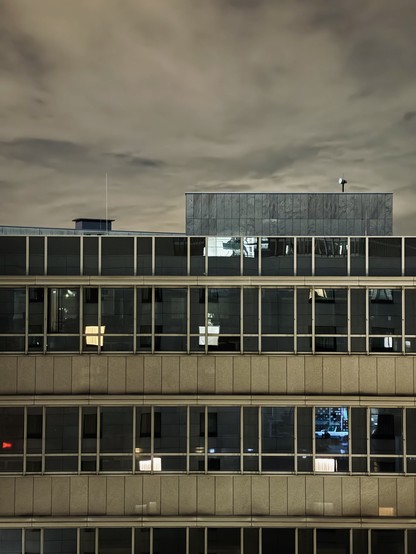 Abends, Blick auf die dunklen oberen Stockwerke eines Bürogebäudes, vereinzelt spiegeln sich erleuchtete Fenster. Darüber bewölkter Himmel.