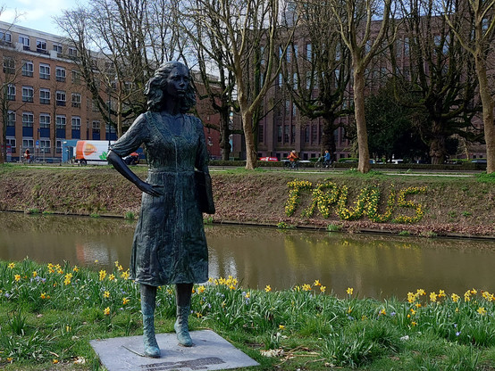 Statue of young woman Truus van Lier before a canal with in the background the name TRUUS in flowering daffodils.