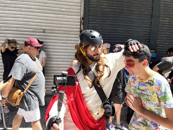 Uma pessoa vestida com um fato semelhante ao de Jesus, com uma faixa vermelha e um capacete, toca na cabeça de um rapaz com sangue falso na cara. Ambos estão rodeados por espectadores num ambiente exterior, e a pessoa fantasiada segura uma câmera.