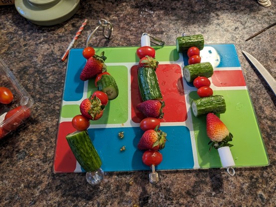3 kabobs on a multicolor cutting board against a granite counter top. The kabob on the left is corkscrew shaped. They each have varying arrangements of mini cucumbers, strawberries, and tomatoes.