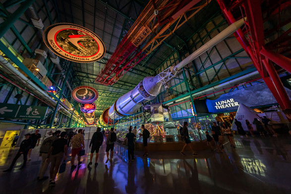A photograph looking down along the Saturn V on display at Kennedy Space Center, from the top of the rocket. Apollo mission logos hang from the ceiling to its left, with the Apollo 15 logo frontmost in the frame and working backwards. The "Lunar Theater" and a capsule are visible on the right, and multiple people visiting the exhibit walk underneath the rocket. 